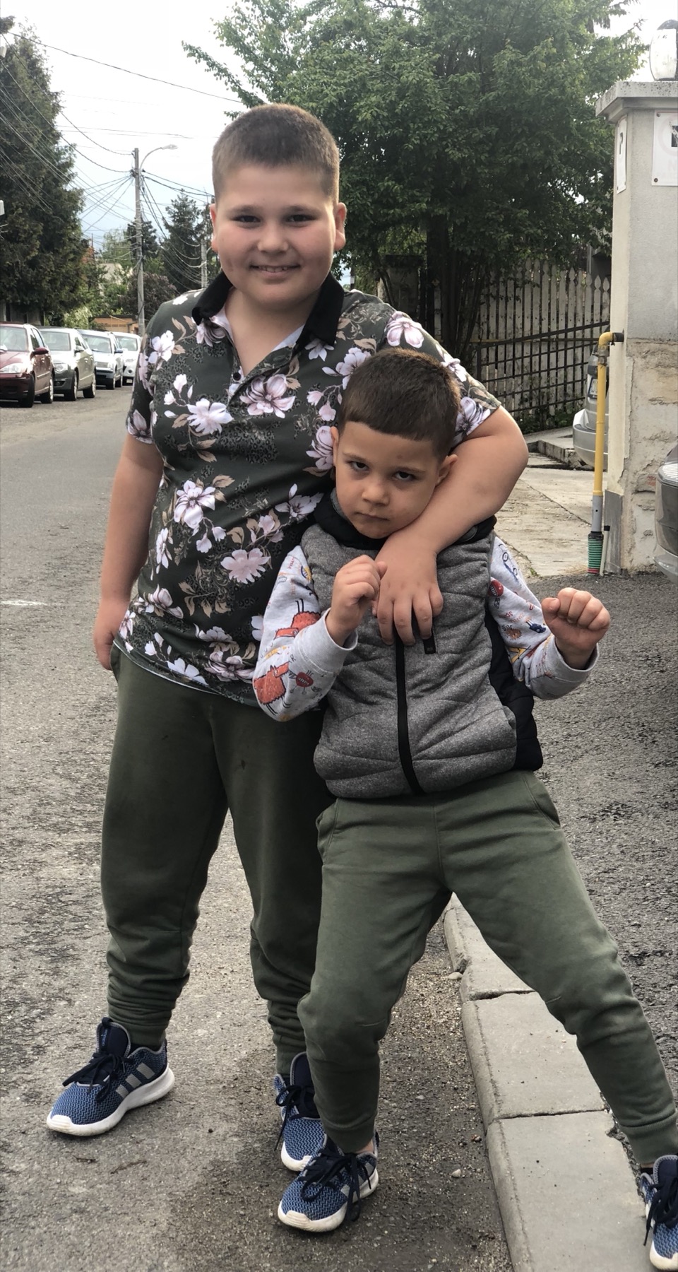Two young boys standing together outside, the older one with his arm around the younger