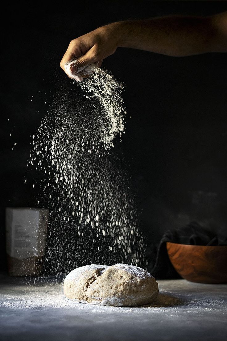 A hand scattering flour over a round of dough, lit against a dark background