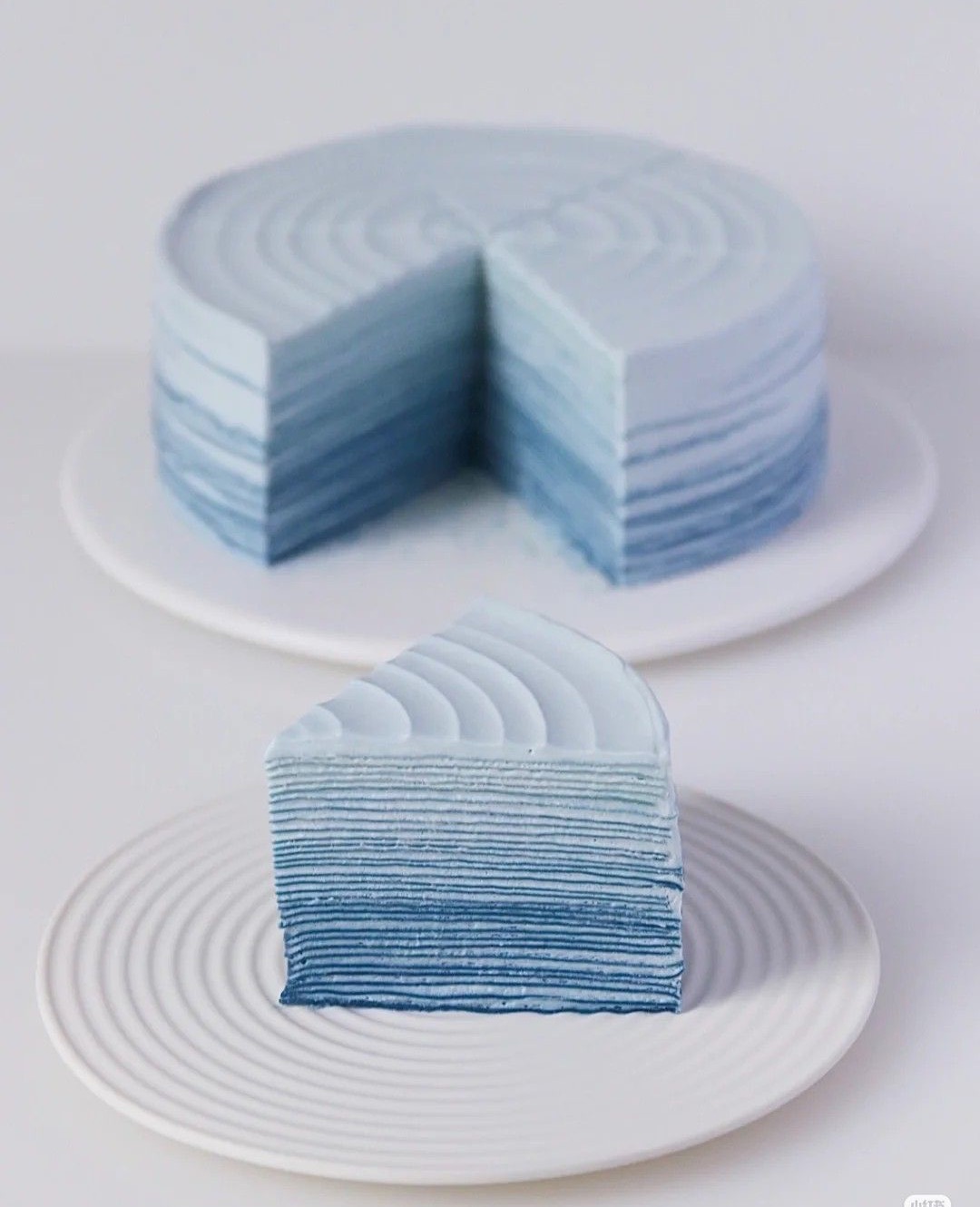 A many-layered crêpe cake in graduated blue tones, one slice cut and plated