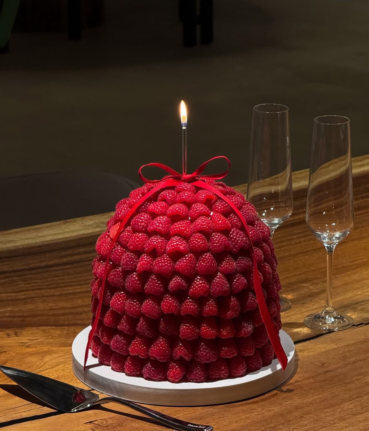 A domed cake covered entirely in fresh raspberries, tied with a red ribbon, one lit candle